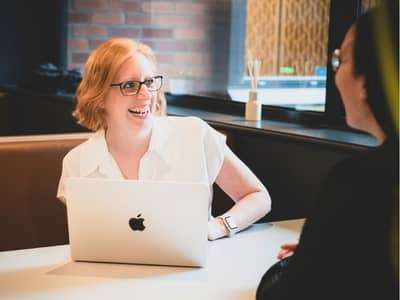 A woman with red hair and glasses sitting at a table smiling at another woman sitting opposite. She is Lucy Davies, business breaththrough coach.